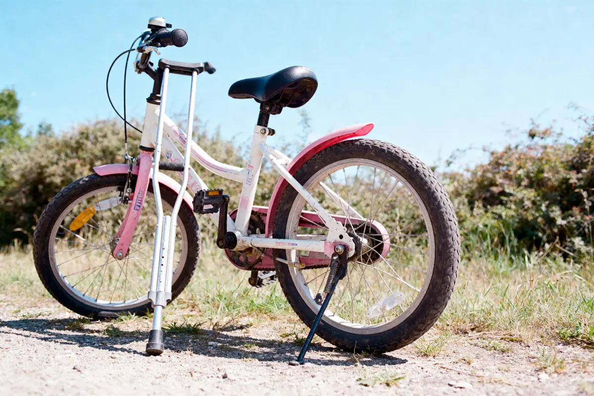 Vélo d'enfant rose et blanc, béquille déployée, posé sur un chemin, ciel bleu en arrière-plan.