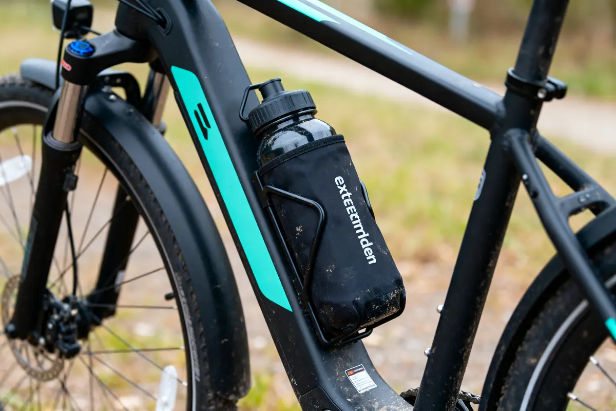 Cadre de vélo noir avec bouteille d'eau noire attachée, logo visible, chemin terreux en arrière-plan.