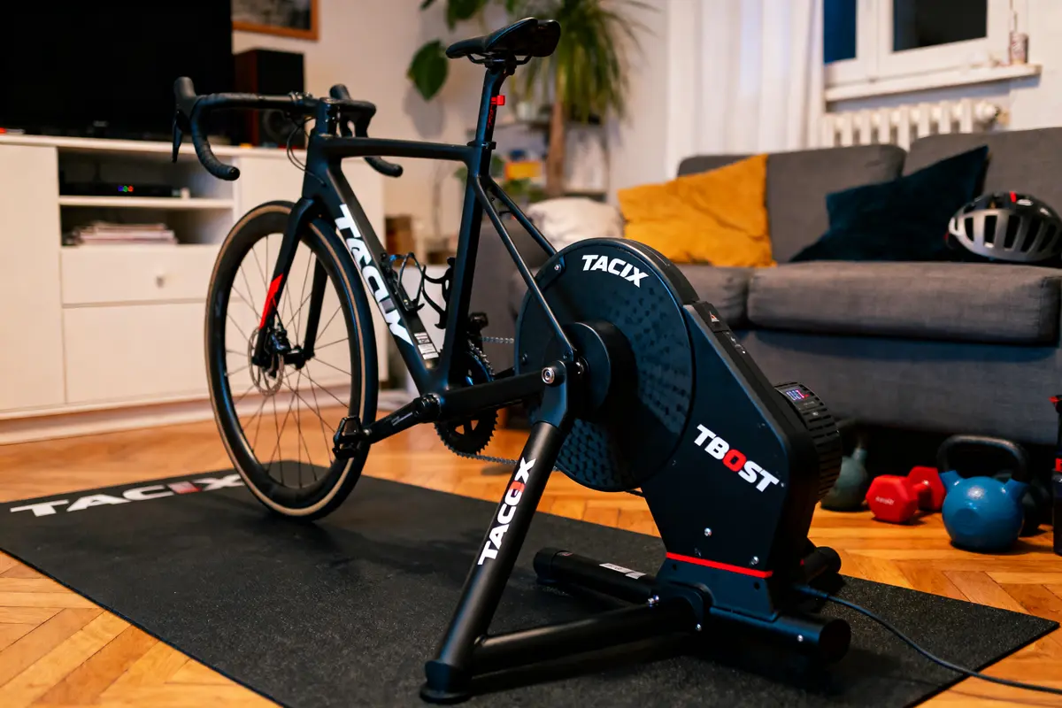 Vélo sur home trainer Tacx dans salon, tapis de sol, canapé gris, haltères colorés en arrière-plan.
